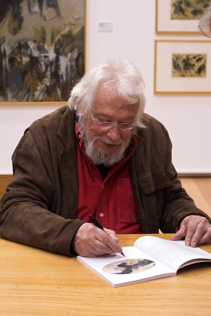 Júlio Pomar sentado a autografar um catálogo. Ao fundo, obras de Pomar expostas na parede.