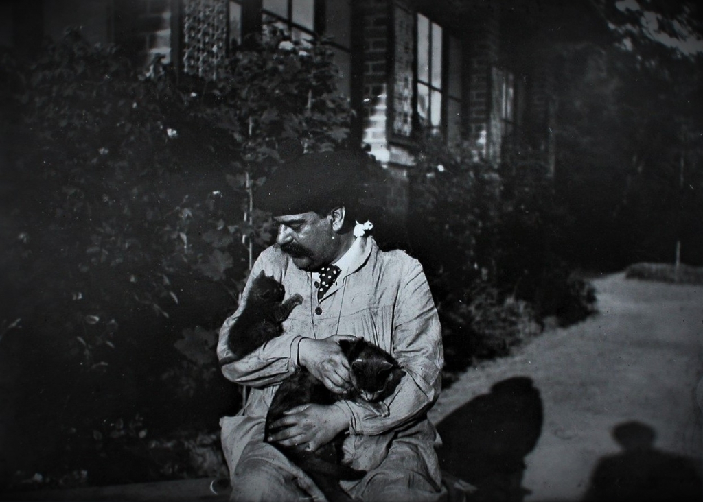 fotografia de um homem com gatos ao colo, sentado em frente a uma casa
