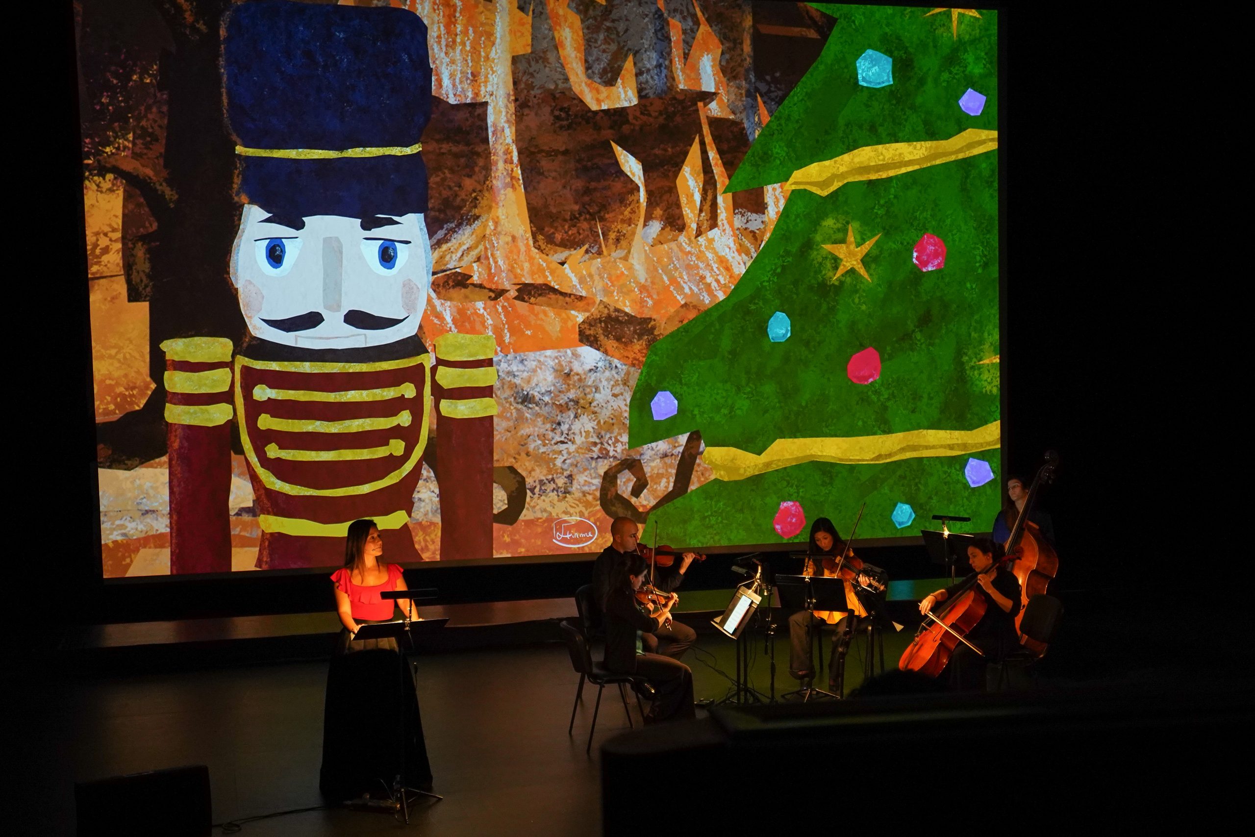 Fotografia. Em palco, músicos sentados e uma mulher, de pé. Em segundo plano, a imagem de um soldadinho e uma árvore com enfeites de natal.
