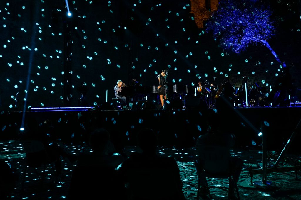 Fotografia noturna. No centro da imagem, um homem a tocar piano, num cenário pontuado por pequenas luzes brilhantes