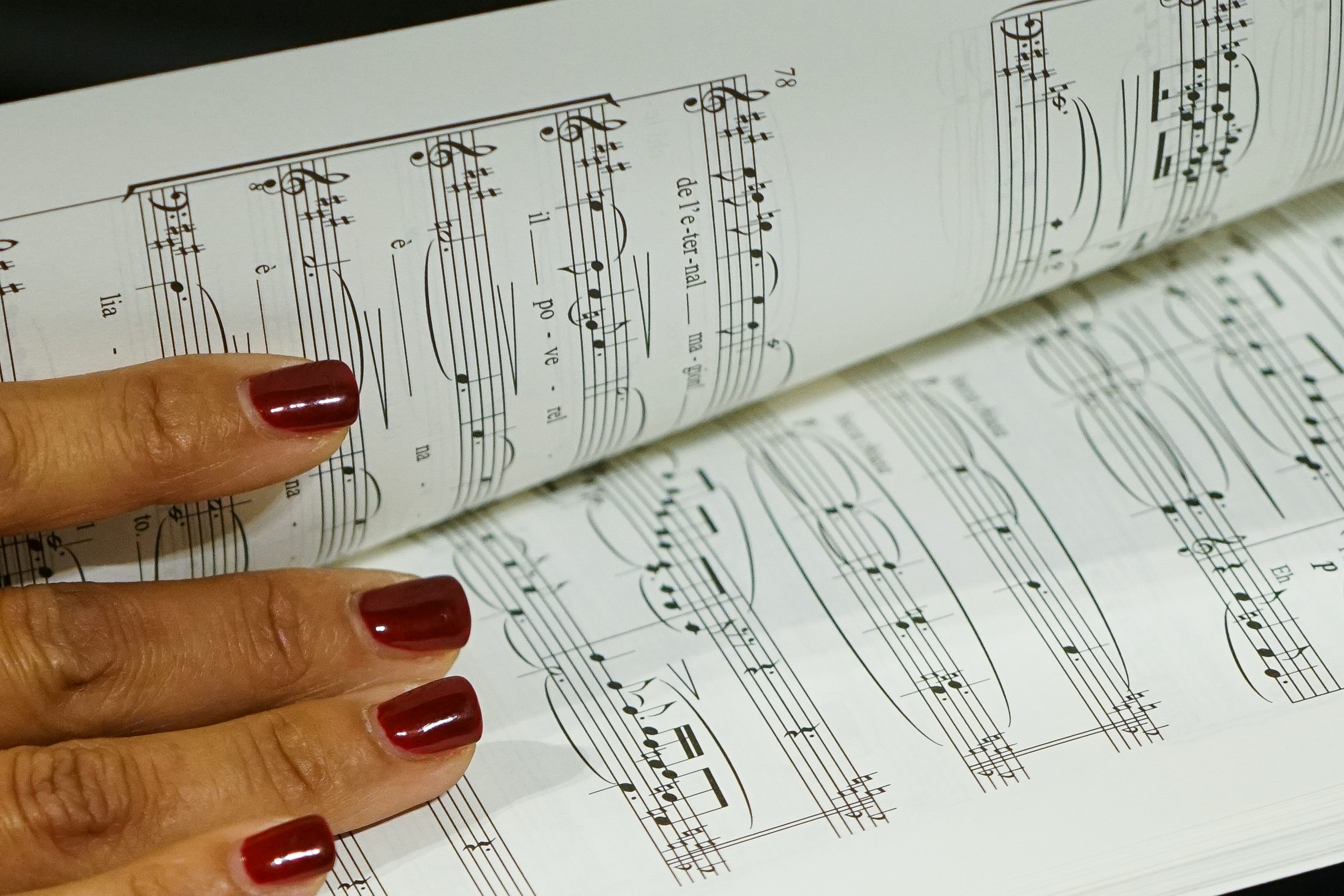 Photography. Detail of musical score. On top is a hand, with nails painted dark red.