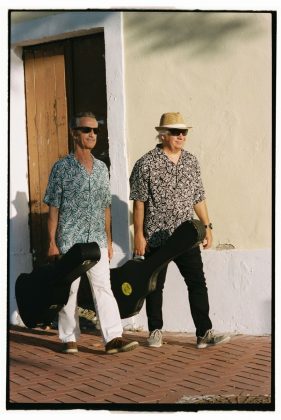 The two band members walk side to side, carrying their guitar boxes, wearing sun glasses and flower shirts