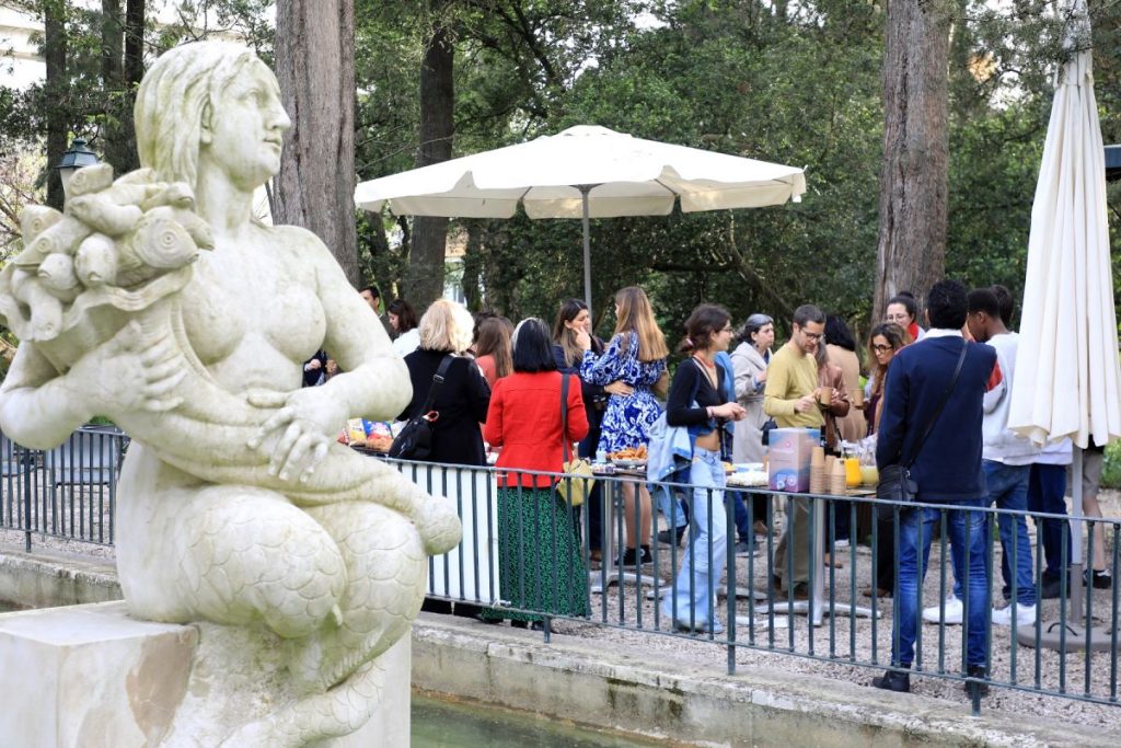 Estátua de sereia no primeiro plano e esplanada com pessoas ao fundo.