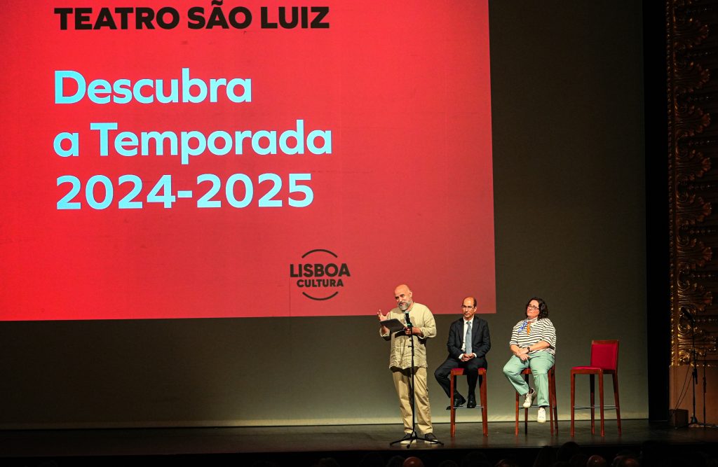 3 pessoas em palco. Na tela de fundo texto 