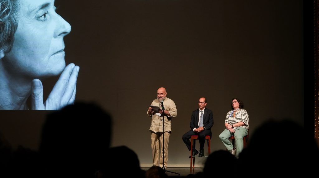 3 pessoas em palco. Na tela de fundo perfil de uma mulher de meia idade