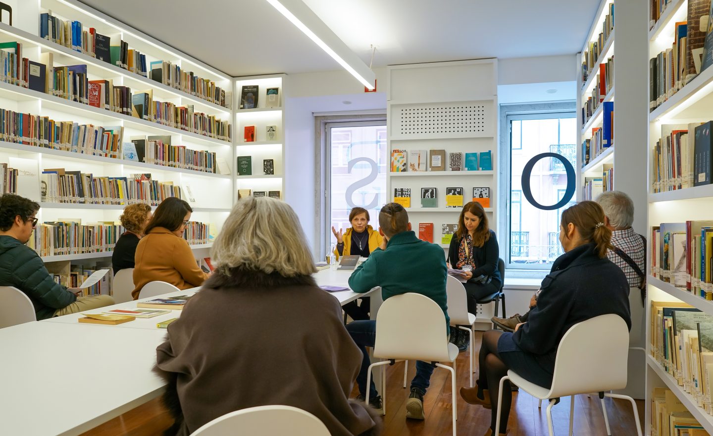 pessoas sentadas em mesas com atenção a 1 mulher a falar, numa biblioteca