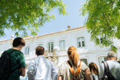 Numa perspectiva de baixo para cima, em primeiro plano, vemos um grupo de pessoas de costas e, em segundo plano, a fachada de um prédio sob um céu azul.