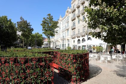 Praça com um jardim de buxo em primeiro plano, com um gradeamento vermelho; algumas árvores e fachadas de edifícios de cinco andares de cor clara