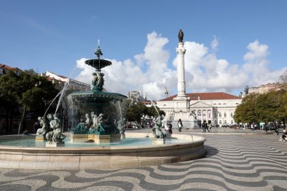 Praça ampla, com uma fonte em primeiro plano à direita e uma estátua num alto pedestal; chão pavimentado em calçada portuguesa recriando as ondas do mar a preto e branco