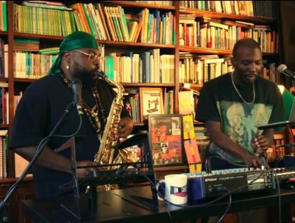 Two African musicians, Kenny Caetano on saxophone and Luzingo Monimambo on keyboard, in the foreground, with a wall of books in the background