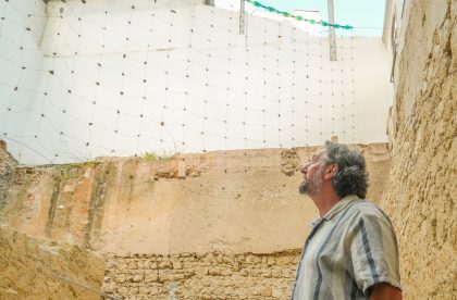 Fotografia a cores onde se vêm paredes de pedra amarela e branca em fundo, em frente das quais está montada uma espécie de rede. Em primeiro plano, do lado direito em baixo, um homem de cabelo grisalho olha para a rede