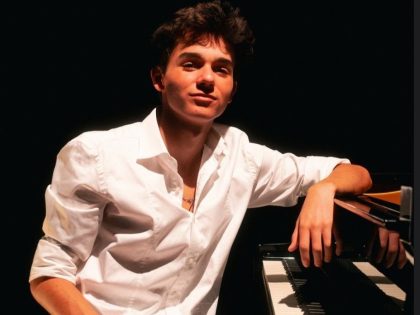 Jovem rapaz, a meio corpo, de camisa branca, sentado em frente a um piano, com o braço esquerdo sobre o piano, num fundo escuro