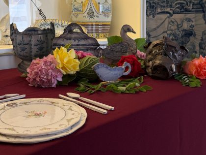 Fotografia a cores, com uma mesa coberta com toalha de pano bordeaux. Em cima da mesa, várias peças a imitar a cerâmica, entre as quais uma cabeça de javali, e flores.