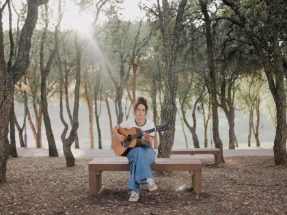 Several trees in the background, with the sun breaking through; in the centre, a young girl sitting on a wooden bench, holding a guitar