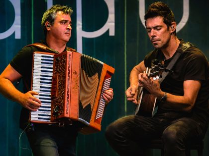 Dois homens sentados, o da esquerda a tocar acordeão e o da direita a tocar viola